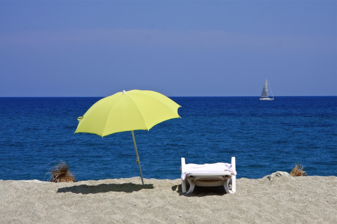 Leerer Liegestuhl mit Sonnenschirm am Strand, im Hintergrund ein Segelboot- erinnert an den wichtigen Schutz der haut vor Sonne.