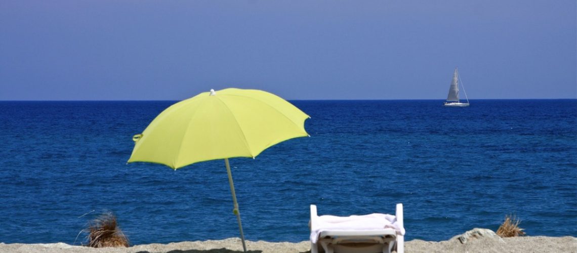 Leerer Liegestuhl mit Sonnenschirm am Strand, im Hintergrund ein Segelboot- erinnert an den wichtigen Schutz der haut vor Sonne.